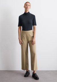 Juodas, trumpomis rankovėmis turtleneko bodysuitas derinamas su smėlio spalvos plačiomis kelnėmis. Modelis vilki juodus platforminius loafers batus. Minimalistinis dizainas, lygus paviršius.