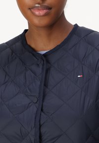 Marineblau gesteppte Jacke mit rundem Kragen und Druckknöpfen. Verfügt über ein kleines Logo in der Nähe des Ausschnitts und eine glatte, strukturierte Oberfläche.