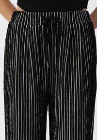 Pantalons noirs avec des rayures verticales blanches, taille élastique et cordon de serrage noir, portés avec un haut noir. Mains sur les hanches.
