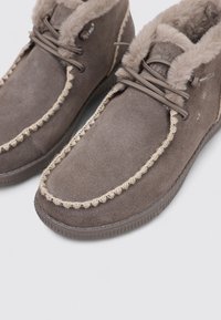 Botas de tobillo de ante marrón con forro de piel sintética, costuras beige en contraste y cierre con cordones. Suela texturizada para mayor agarre y comodidad.