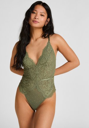 Žena s dlhými tmavými vlasmi, oblečená v olivovo zelenej čipkovanej body s tenkými ramienkami, stojaca na jednoduchom svetlosivom pozadí.