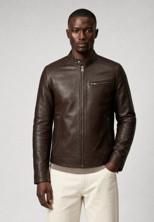 Man draagt een donkerbruine leren jas met ritszakken en beige broek, staand tegen een effen lichtgrijze achtergrond.