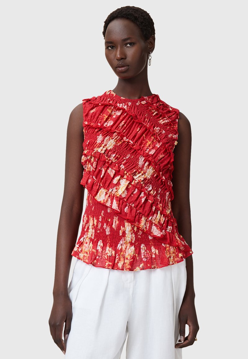 Blusa rossa senza maniche con texture a cerchi, caratterizzata da un motivo floreale giallo e arancione. Leggermente aderente in vita, abbinata a pantaloni bianchi.