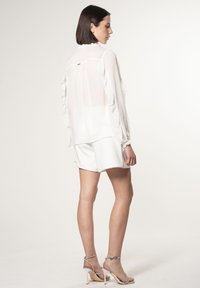 Blusa bianca trasparente con maniche a volant, abbinata a pantaloni corti bianchi. Presenta una chiusura con bottoni sul retro e tacchi a strisce argentati.