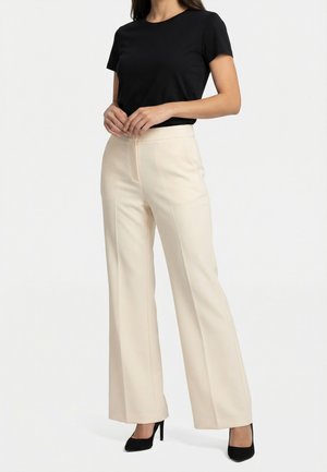 Mujer con blusa negra de manga corta, pantalones anchos beige y zapatos de tacón negros, de pie con las manos entrelazadas a la altura de la cintura.