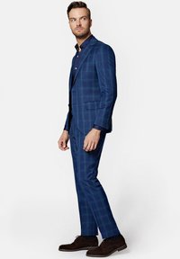 Granatowy garnitur w kratę o slim fit, z klapami w szpic i dopasowaną niebieską koszulą. Brązowe buty dopełniają stylizację.