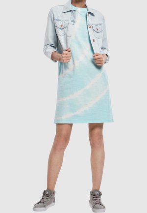 Osoba ubrana w jasnoniebieską sukienkę w stylu tie-dye z jasną dżinsową kurtką i szarymi sneakersami typu high-top, stojąca na tle jednolitym.