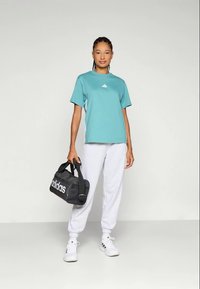 Türkiissinine Adidas T-särk, valge logo, hallide dressipükste ja valgete tossudega, hoides musta spordikotti valge brändinguga.