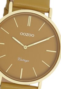 Montre marron avec un cadran à finition mate, index et aiguilles dorés, montée sur un bracelet en cuir texturé couleur beige. Présente "OOZOO Vintage."