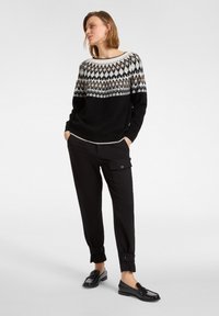 Kobieta ubrana w wzorzysty sweter w kolorach czarnym, białym i brązowym, czarne spodnie cargo z mankietami na guziki oraz czarne mokasyny, stojąca z rękami w kieszeniach.