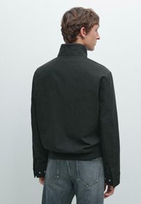 Black, collared jacket made of textured fabric, featuring a relaxed fit, back yoke, and buttoned cuffs, paired with grey denim.