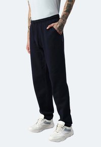 Marineblaue Sweatpants mit tapered Fit, elastischem Bund, Seitentaschen und weichem Material; kombiniert mit weißen Sneakers mit grauen Akzenten.