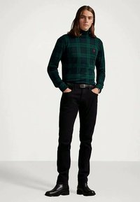 Fiatal férfi vállig érő hajjal, sötétzöld és fekete kockás garbó pulcsiban, fekete nadrágban és fekete csizmában áll, kezét zsebébe dugva.