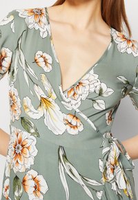 Kukallinen wrap-mekko hillityssä vihreässä kankaassa, jossa on valkoisia, oransseja ja keltaisia kukkia; syvä V-pääntie ja sivusolkiyksityiskohta.