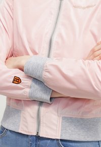 Personne portant une veste bomber rose pâle avec des poignets et une ceinture gris, bras croisés, vue partielle de la taille aux épaules.