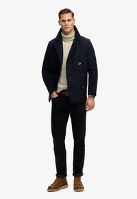 Man draagt een marineblauwe pea coat, een beige coltrui, zwarte broek, bruine riem, bruine laarzen en een polshorloge, en staat met één hand in zijn zak.