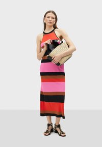 Striped knit dress in pink, red, and black. The model holds a woven bag, wearing black studded sandals. Smooth texture, form-fitting design.