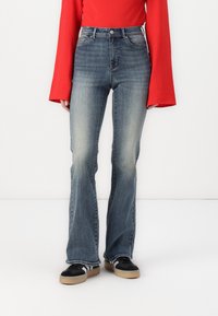 Mørkeblå flarejeans med en falmet vask, med høy midje og kontraststing, parret med svarte sneakers og en rød langermet topp.