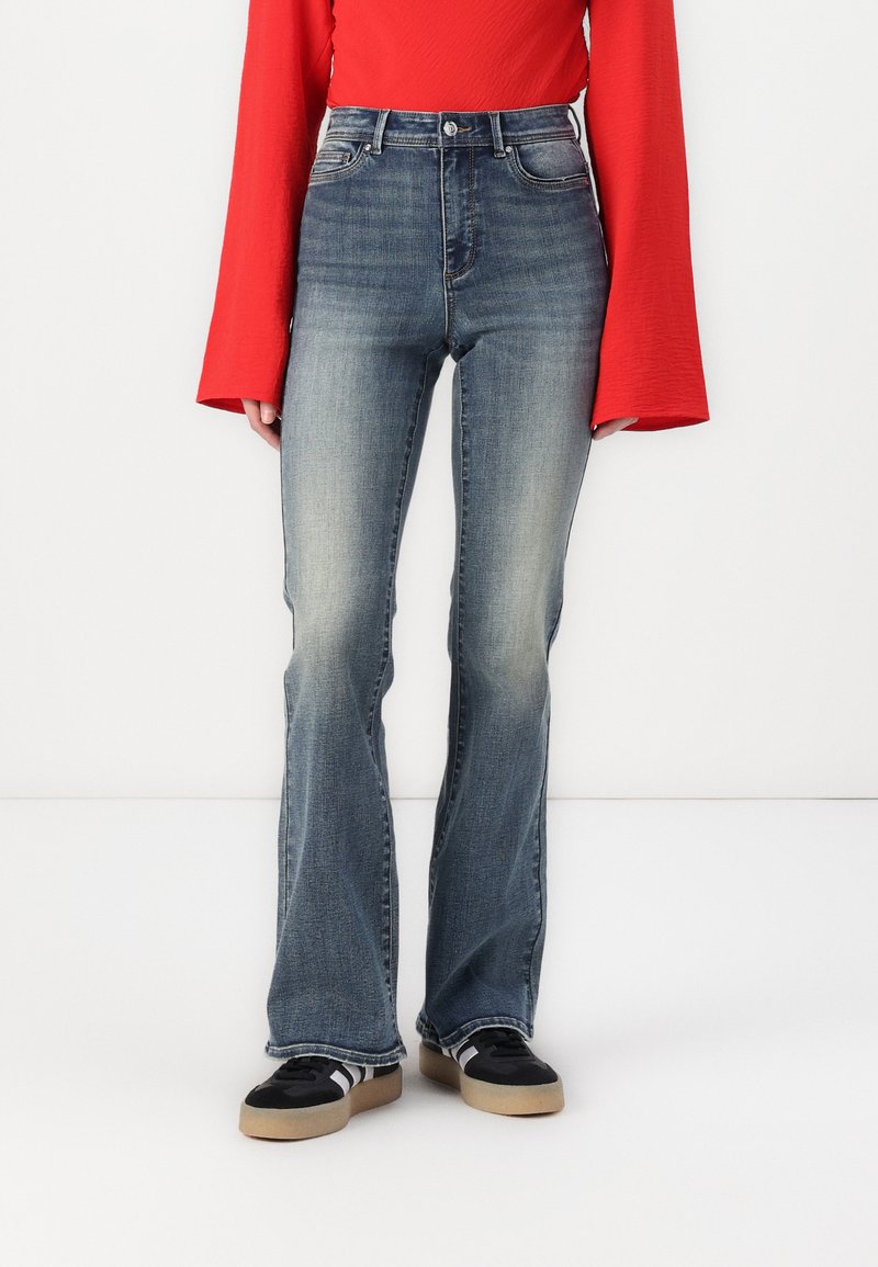 Mørkeblå flarejeans med en falmet vask, med høy midje og kontraststing, parret med svarte sneakers og en rød langermet topp.