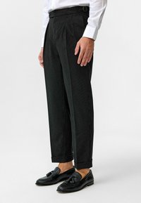 Zwarte pinstripe broek met een getailleerde pasvorm, plooien aan de voorkant en riemlussen. Draag met zwarte instappers, wat een formele stijl toont.