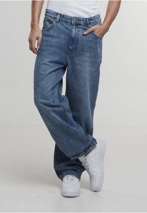 Alsótest laza, kék farmert és fehér tornacipőt visel, egyik kezével zsebében, lábait keresztbe téve, egyszínű háttér előtt.