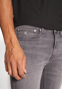 Jeans de denim gris con un ajuste slim, que presentan un pequeño logo en el bolsillo frontal, diseño estándar de cinco bolsillos y detalles en botón metálico.