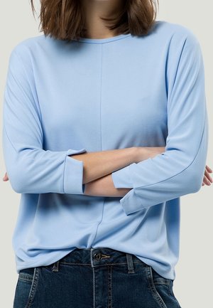 Světle modrý dlouhý rukáv top z hladké látky, s kulatým výstřihem a otočenými manžetami, nošený s tmavými džínami.