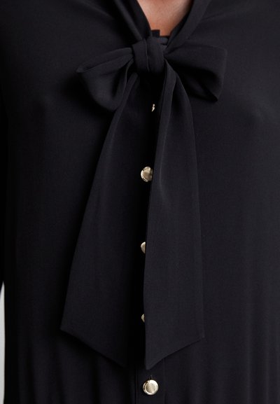Blouse noire avec un nœud de cou noué, texture lisse et boutons métalliques à l'avant pour la fermeture.