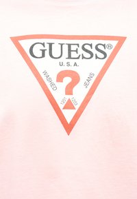 Κόκκινο τρίγωνο λογότυπο με τη φράση «GUESS U.S.A.», ένα μεγάλο ερωτηματικό και τις λέξεις «WASHED» και «JEANS» σε ελαφρώς ροζ ύφασμα.
