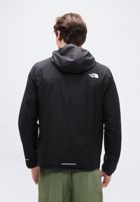 Άνδρας που φοράει μαύρο μπουφάν με κουκούλα The North Face και πράσινο παντελόνι, βλεπόμενος από πίσω σε απλό φωτεινό φόντο.