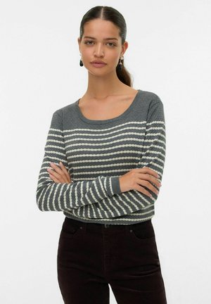 Pull en tricot gris avec des rayures horizontales blanches, col rond et manches longues. Associé à un pantalon en velours côtelé foncé.