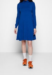 Blå långärmad klänning med hög krage, avslappnad passform och utsvängd fåll. Matchas med orangea sportskor och vita strumpor.