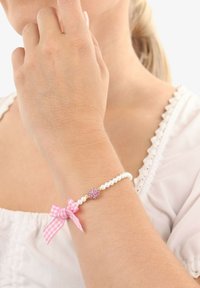 Armband med vita pärlor, ett charm i form av ett pink glittrande hjärta och en rosa gingham-bow, bärs på handleden, hand som rör vid ansiktet, vit klädbakgrund.