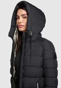 Doudoune noire avec une grande capuche, un design matelassé et une doublure métallique. Comprend un écusson de marque sur la manche et un cordon de serrage.