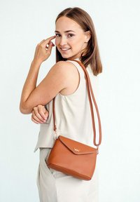 Hnědá kožená crossbody taška s klopou, řetízkovým zdobením a nastavitelného popruhem. Umístěná na světlém pozadí.