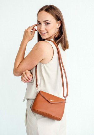 Hnědá kožená crossbody taška s klopou, řetízkovým zdobením a nastavitelného popruhem. Umístěná na světlém pozadí.
