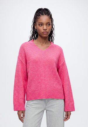 Jeune femme aux cheveux tressés portant un pull rose vif à col en V et un pantalon gris clair, debout devant un fond blanc uni.
