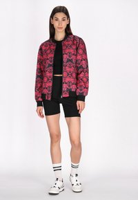 Rood en zwart gestreepte bomberjack met zwarte afwerking, gecombineerd met een zwarte crop top en zwarte fietsbroek, compleet met witte sneakers.