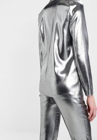 Metalický stříbrný oblek s upraveným sakem a přiléhavými kalhotami. Hladký povrch s lesklým finišem, který ukazuje elegantní siluetu.