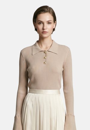 Donna con capelli corti che indossa un maglione polo beige a coste con maniche lunghe e bottoni dorati e una gonna bianca plissettata su uno sfondo semplice.