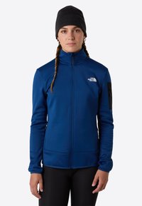 Modrá fleecová bunda s vysokým límcem a zipem. Obsahuje černou kapsu na rukávu a vyšívané logo. Měkká textura a atletický střih.
