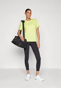 Lysegul atletisk t-shirt, sorte leggings og hvide sneakers. Modellen bærer en sort sportstaske med tekstureret overflade og kontrasterende detaljer.
