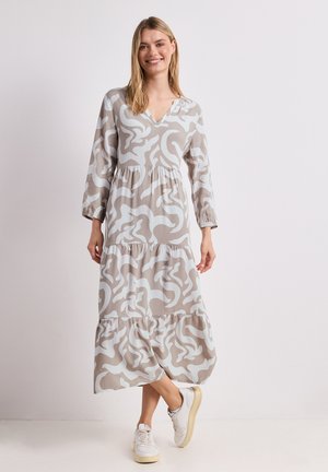 Femme souriante portant une robe beige à manches longues avec col en V, motif abstrait blanc et baskets blanches, debout devant un fond uni.