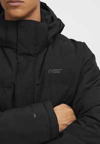 Čierna zateplená bunda s vysokým golierom a nastaviteľnou kapucňou. Vyznačuje sa logom "NORTH BEND" na hrudi a textúrovaným povrchom látky.
