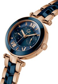 Orologio in tonalità oro con un quadrante lucido di colore blu scuro caratterizzato da numeri romani. Bracciale metallico a maglie con dettagli blu scuro e corona.