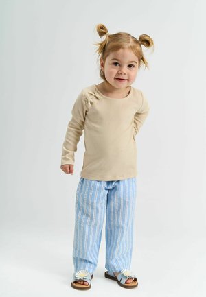 Mała dziewczynka uśmiechająca się z kucykami, ubrana w beżowy sweter z długim rękawem, niebieskie spodenki w paski i sandały z dekoracjami w kształcie stokrotek.