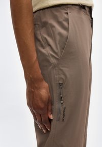 Κοντινό πλάνο ενός χεριού που ακουμπά δίπλα σε μπεζ παντελόνια Salomon με τσέπη με φερμουάρ και ορατές ραφές.