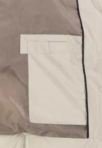 Leichter, beiger Stoff mit strukturierter Oberfläche. Verfügt über eine seitliche Tasche mit Klappe und einem kontrastierenden grauen Panel dahinter. Genähte Kanten.
