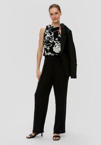 Schwarzes ärmelloses Top mit weißem Blumenmuster, kombiniert mit schwarzen Anzughosen und einem schwarzen Blazer, der über die Schulter drapiert ist. Schwarze Sandalen mit Absatz.