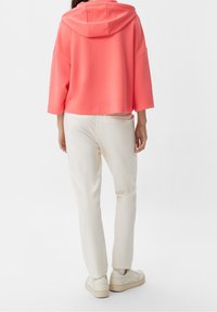 Koralowa, krótka bluza z kapturem, szerokimi rękawami, zestawiona z jasnobeżowymi, taperowanymi spodniami i jasnymi trampkami. Gładka faktura, casualowy design.
